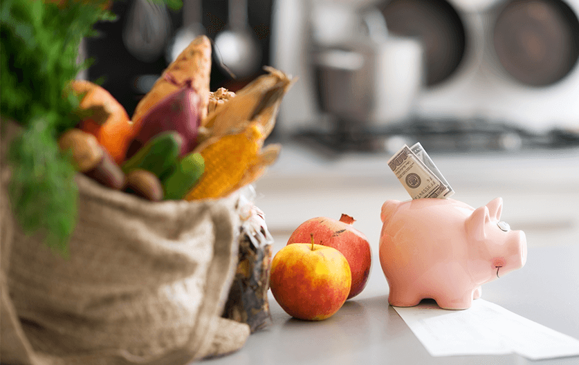 A bag full of healthy groceries next to a piggy bank with money poking out.